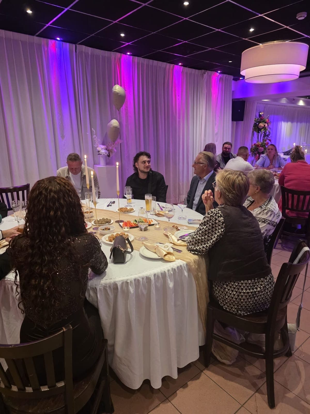 Gasten aan een ronde tafel met witte rok, gouden loper en kaarsen, met hapjes op tafel, onder paars en roze licht
