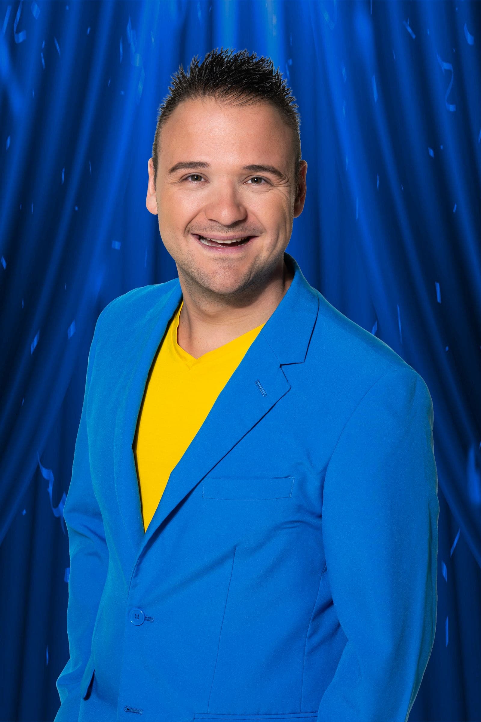 Marco Kraats lachend in zijn blauwe jasje met geel shirt, voor een blauw theaterdoek
