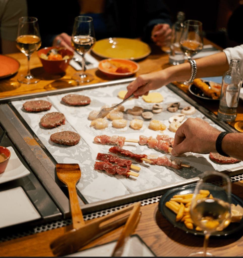 Twee handen leggen biefstukjes, sint-jakobsschelpen en spiesjes op de grillplaat midden in de tafel