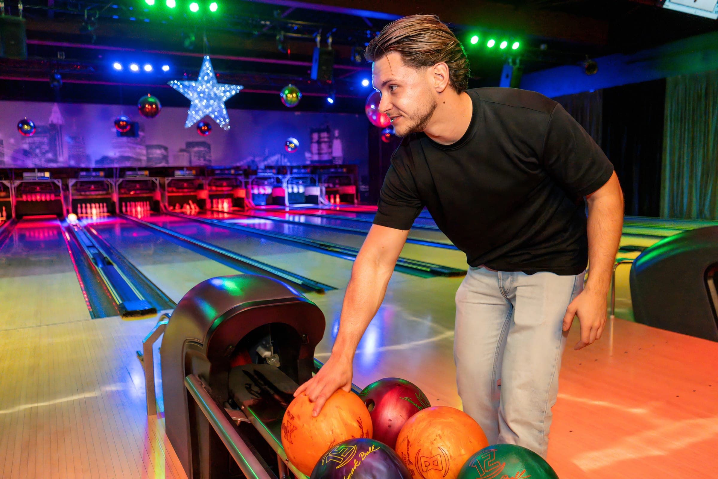 Een gast pakt een bowlingbal uit de ballenretour, met de verlichte banen en lichtsterren achter hem