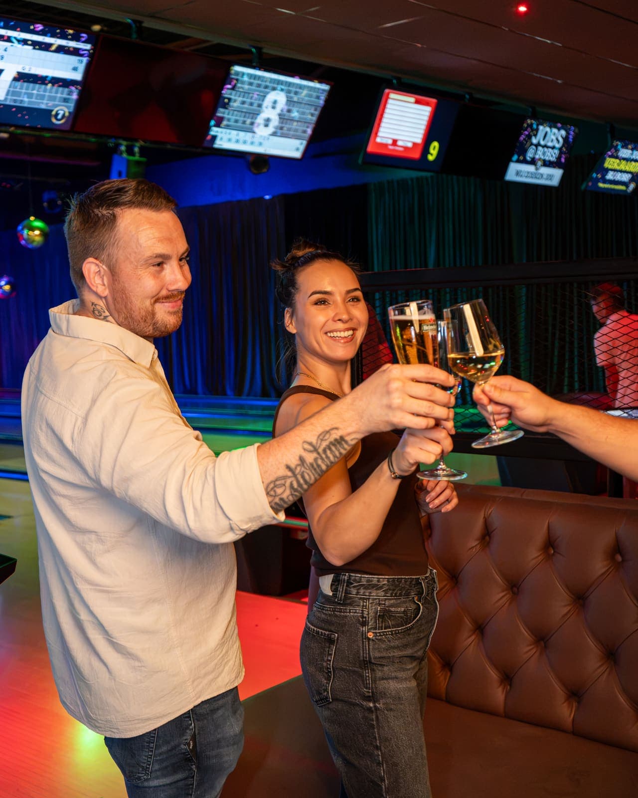 Twee gasten proosten met bier en wijn bij de banen, met de scoreschermen boven hen