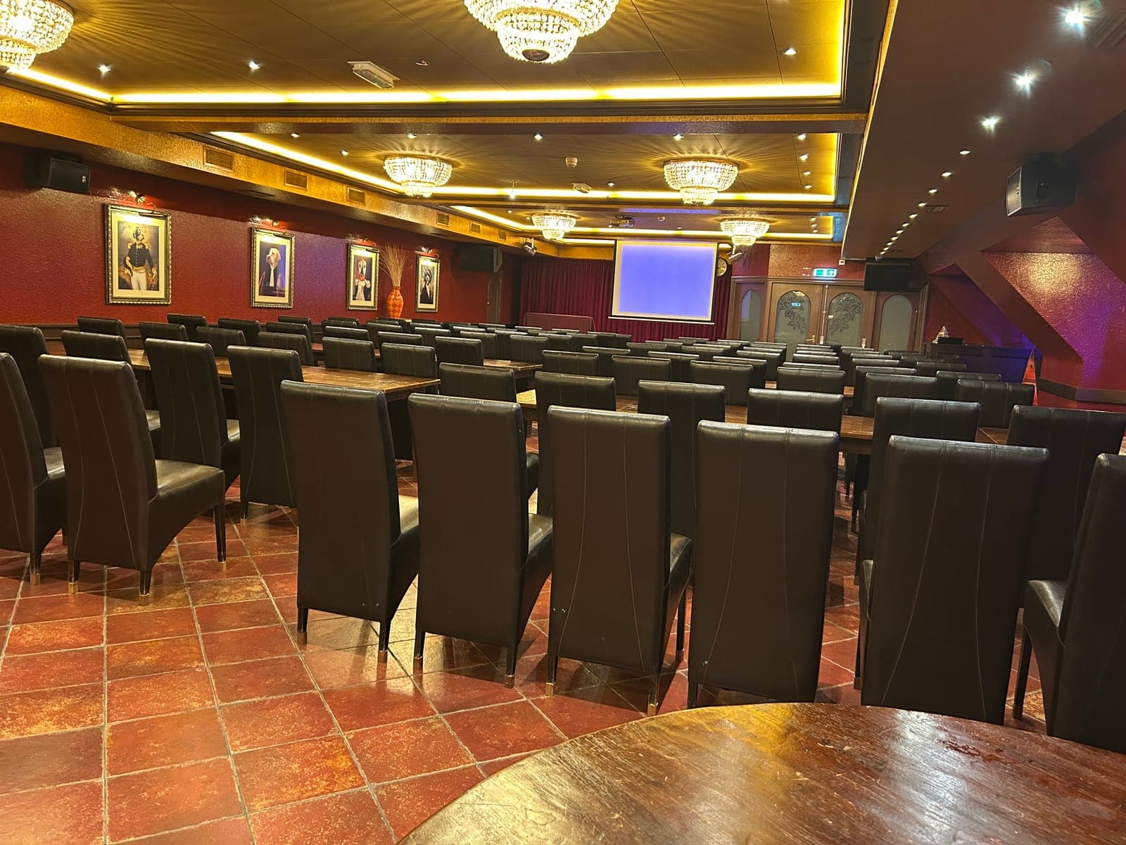 Rijen stoelen gericht op een projectiescherm, in een zaal met kroonluchters